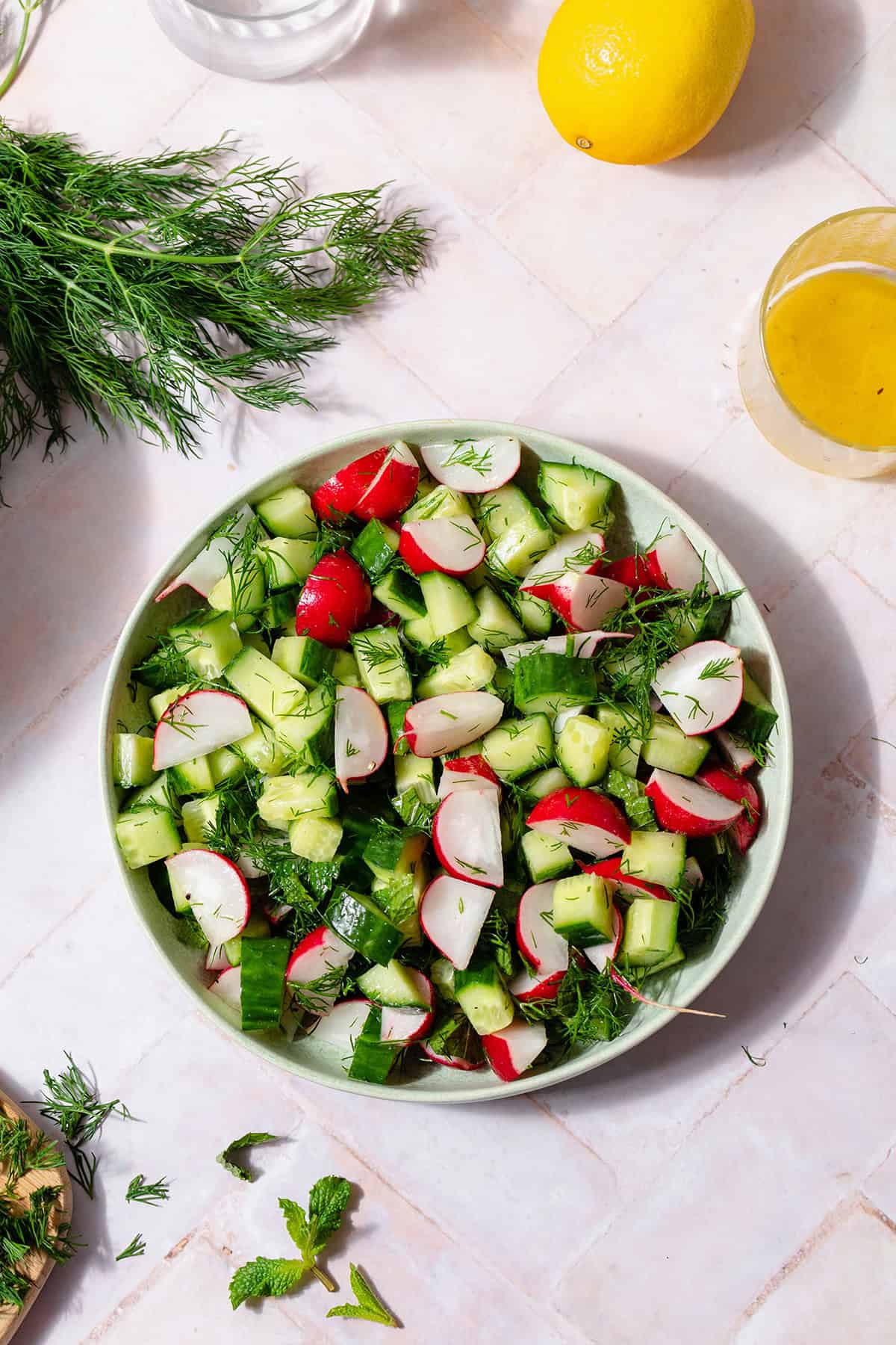 Cucumber Radish Salad in a shallow bowl on a light pink tile background. A bunch of fresh dill on the left side of the plate and the lemon vinaigrette on the right side of it.