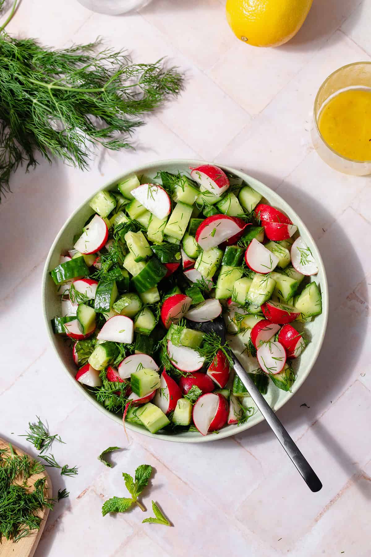 Cucumber Radish Salad in a shallow bowl with a black fork. On a light pink tile background. A bunch of fresh dill on the left side of the plate and the lemon vinaigrette on the right side of it.