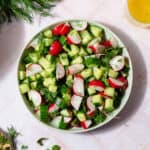 Cucumber Radish Salad in a shallow bowl on a light pink tile background. A bunch of fresh dill on the left side of the plate and the lemon vinaigrette on the right side of it.