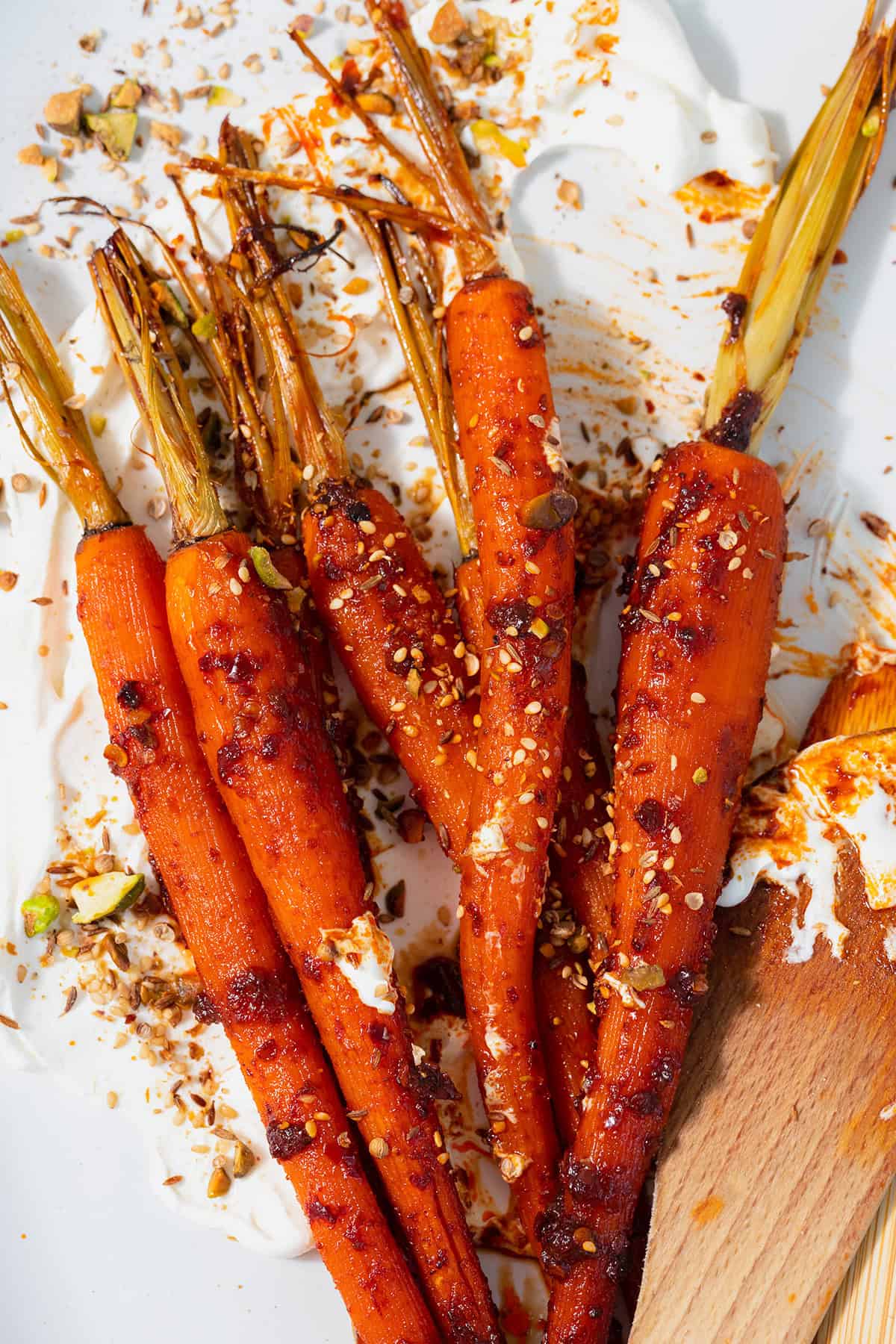 A tight close up of Harissa roasted carrots on a white serving platter with labneh spread under the carrots. Sprinkled with dukkah. Two wooden spoons messy with labneh and harissa leaning on the right edge of the platter. Plate on a white and turquoise tile table.