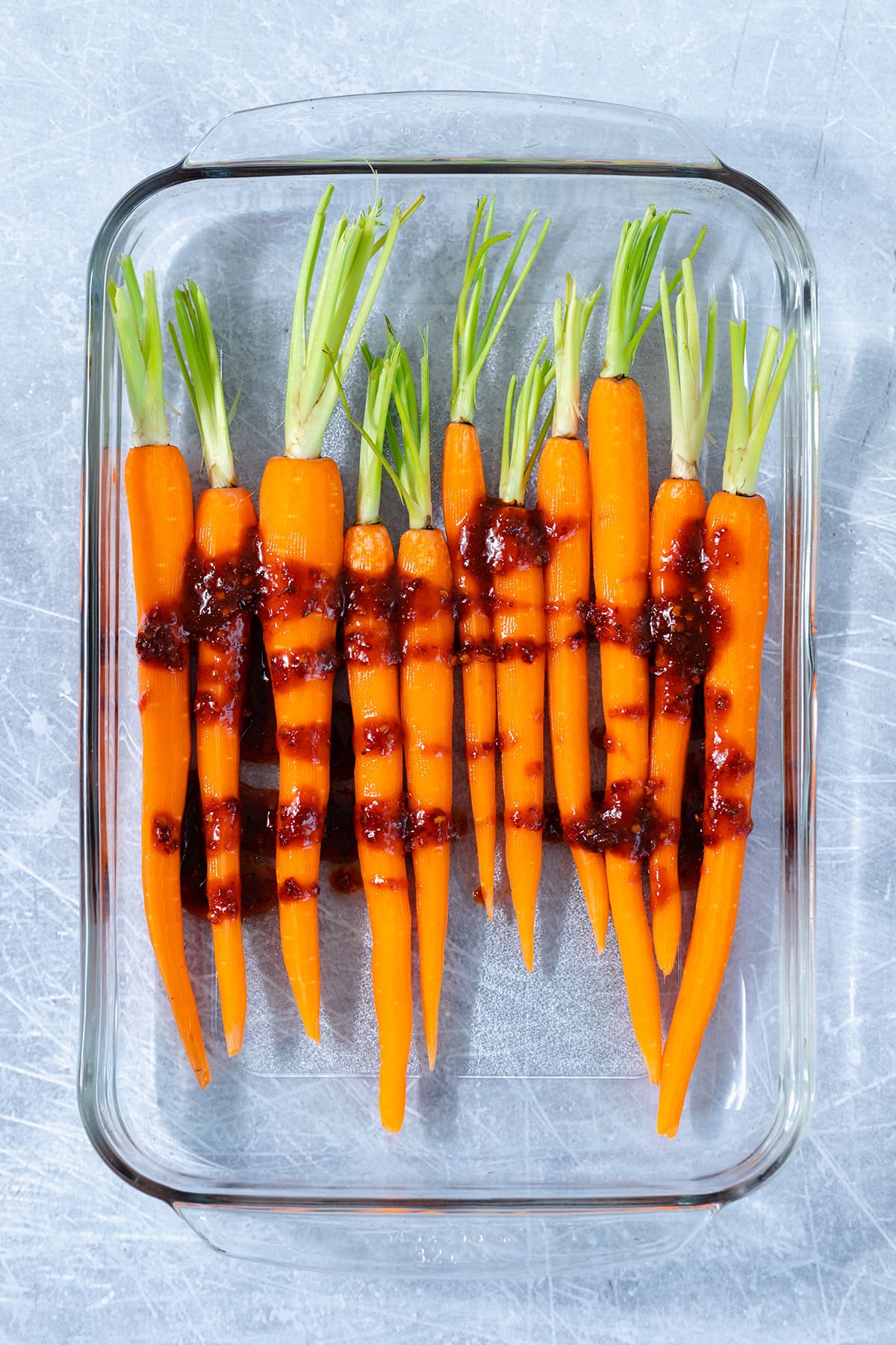 Raw peeled carrots in a glass baking dish with harissa sauce drizzled on them. Dish on a blue scratched metal background.