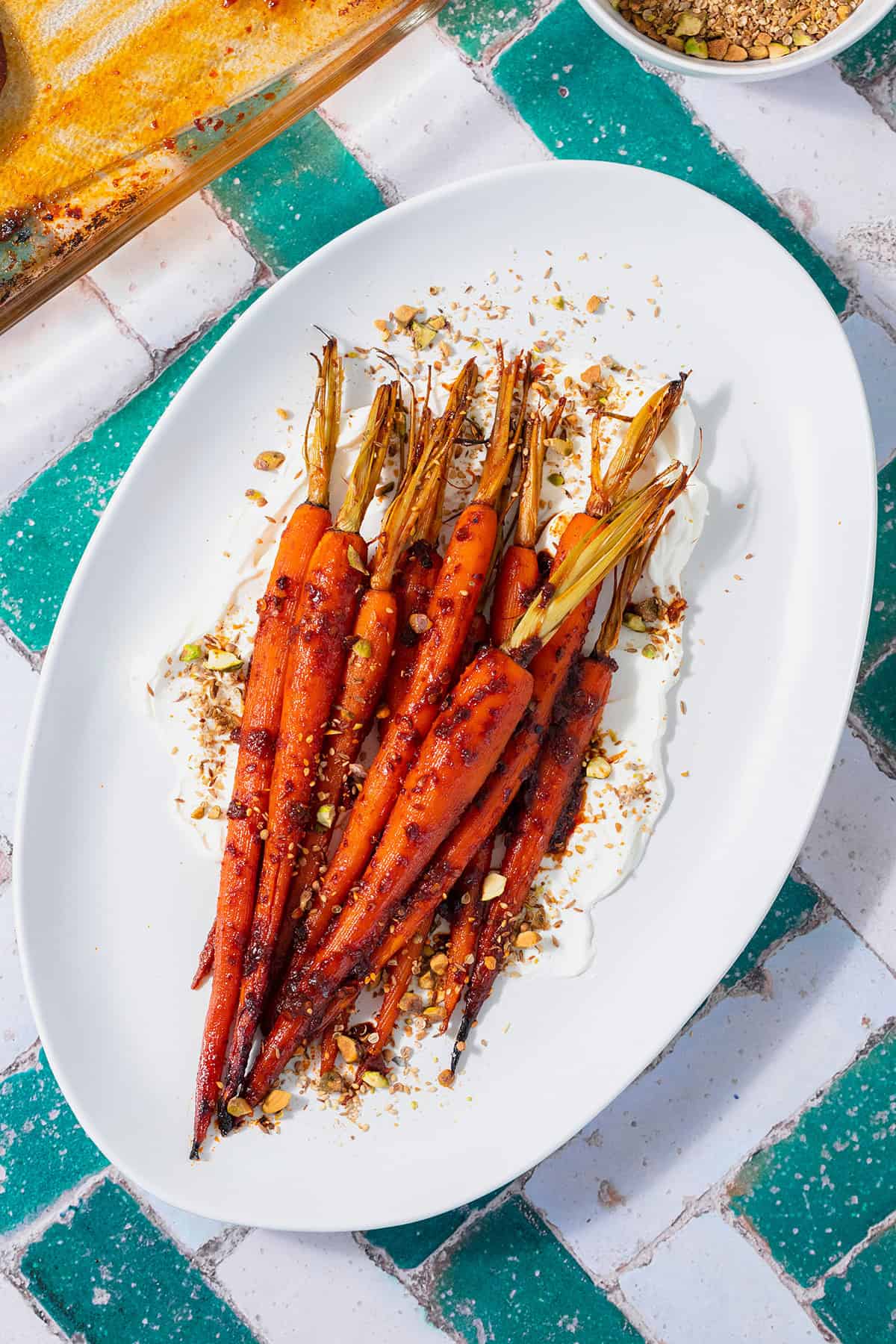 Harissa roasted carrots on a white serving platter with labneh spread under the carrots. Sprinkled with dukkah. Plate on a white and turquoise tile table.