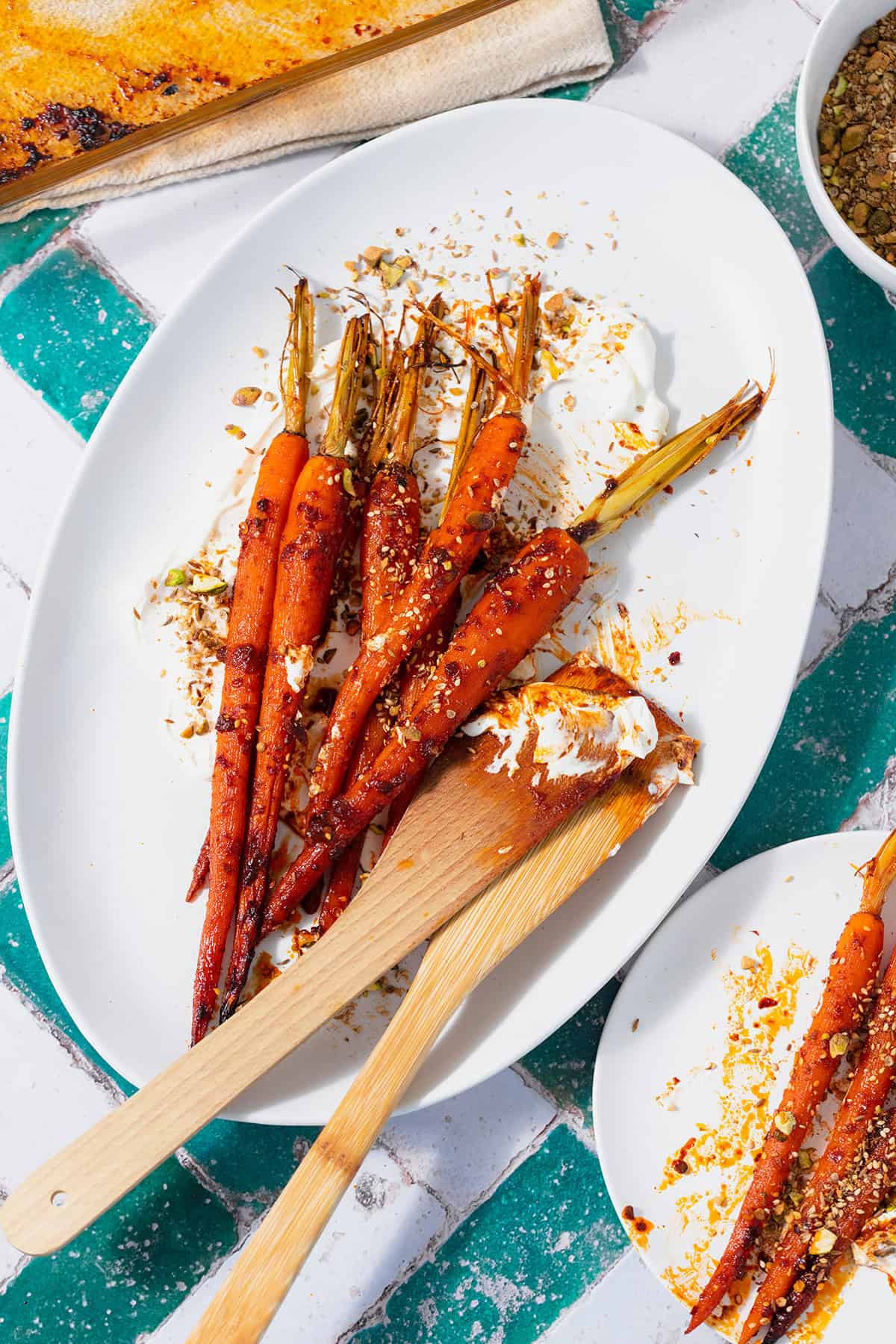 Harissa roasted carrots on a white serving platter with labneh spread under the carrots. Sprinkled with dukkah. Two wooden spoons messy with labneh and harissa leaning on the right edge of the platter. Plate on a white and turquoise tile table.