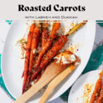 Harissa roasted carrots on a white serving platter with labneh spread under the carrots. Sprinkled with dukkah. Two wooden spoons messy with labneh and harissa leaning on the right edge of the platter. Plate on a white and turquoise tile table.