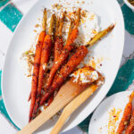 Harissa roasted carrots on a white serving platter with labneh spread under the carrots. Sprinkled with dukkah. Two wooden spoons messy with labneh and harissa leaning on the right edge of the platter. Plate on a white and turquoise tile table.