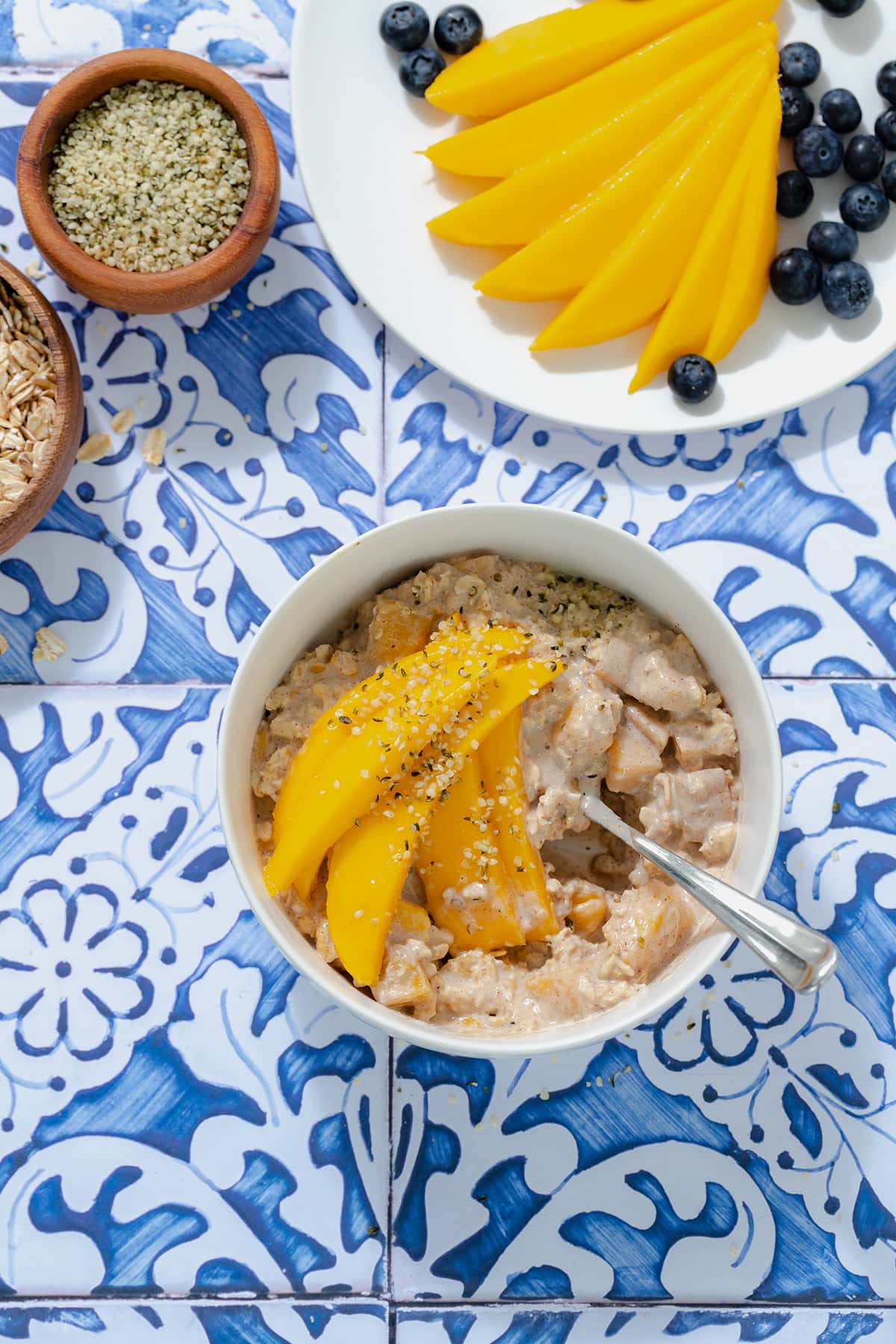 Mango overnight oats in a white bowl on a blue tile background. Mango on a white plate in the top right corner.