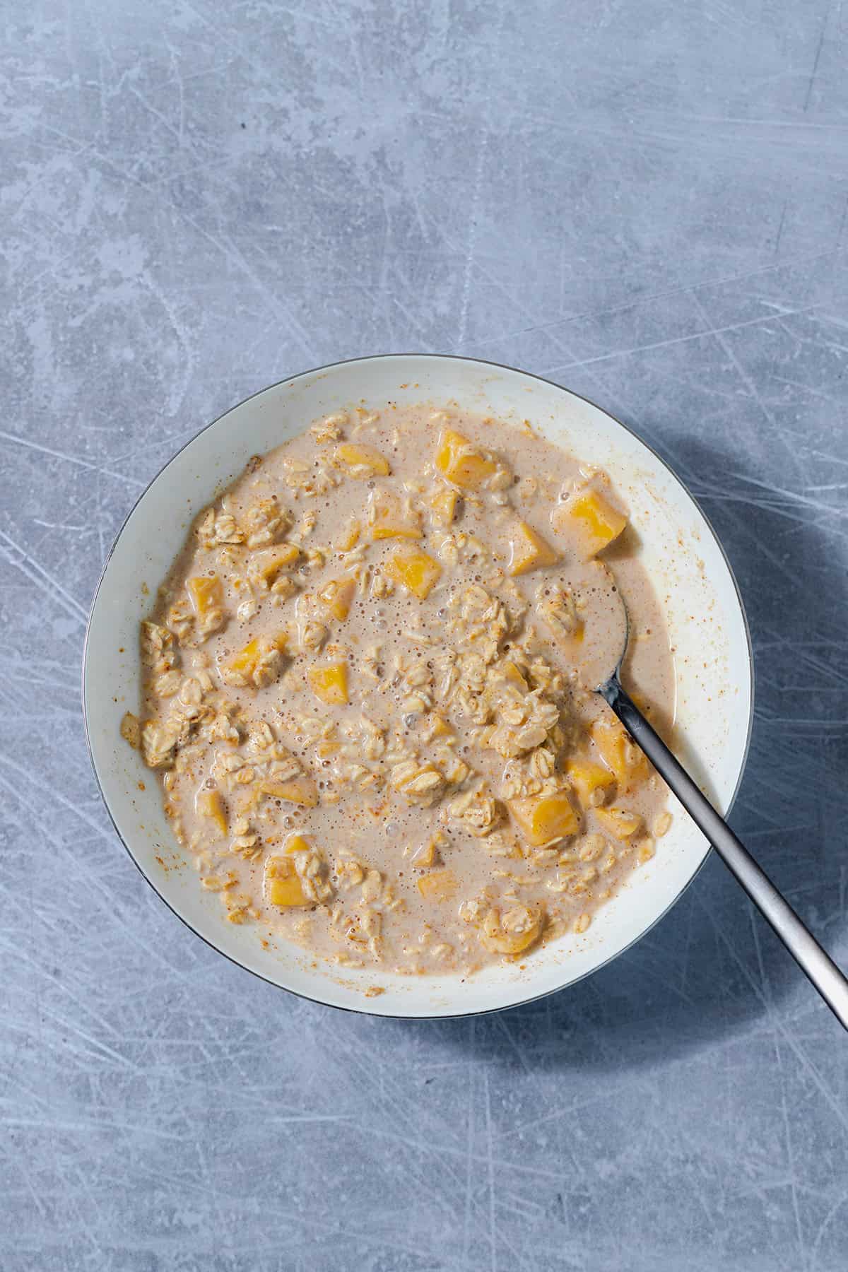 Overnight oats all mixed with with mango in a white shallow bowl on a scratched grey background.