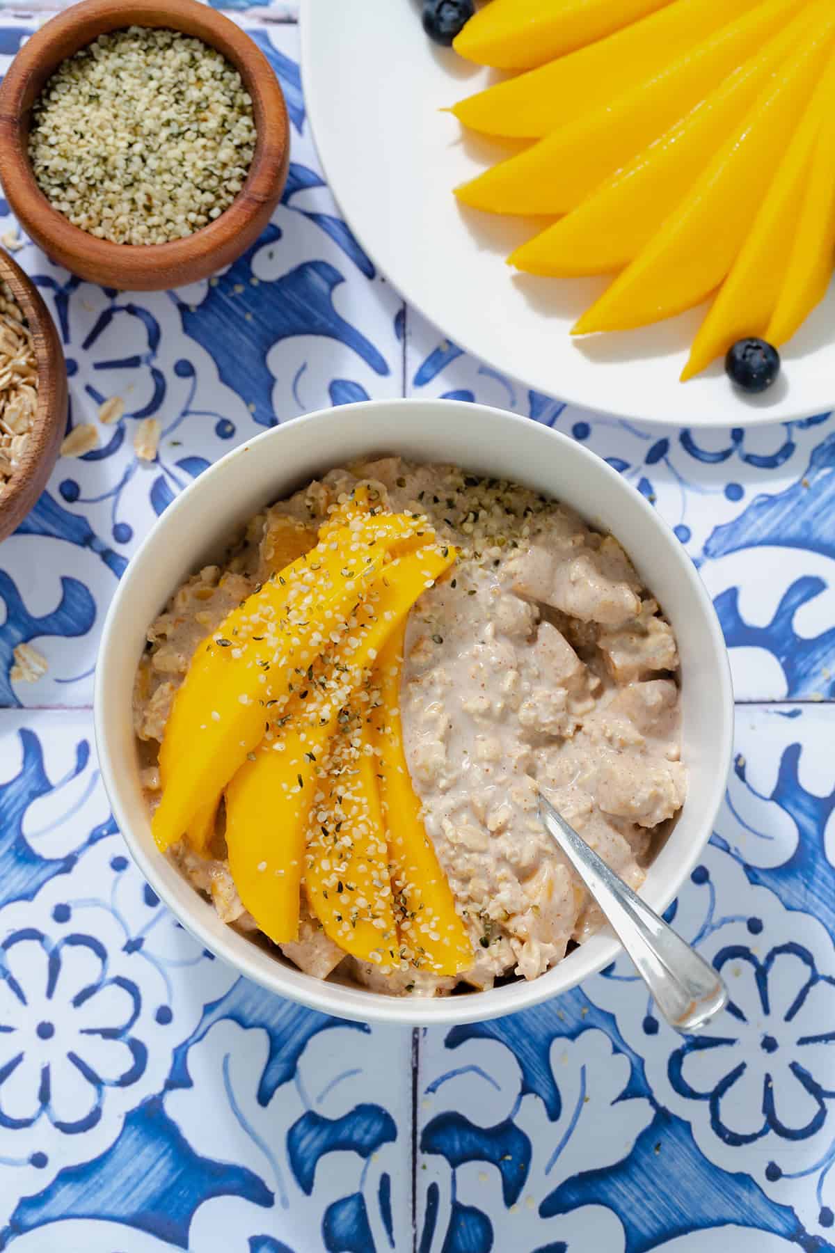 Mango overnight oats in a white bowl on a blue tile background. Mango on a white plate in the top right corner.