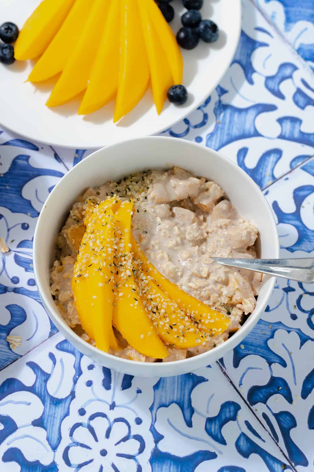 Mango overnight oats in a white bowl on a blue tile background. Mango on a white plate in the top left corner.