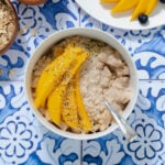 Mango overnight oats in a white bowl on a blue tile background. Mango on a white plate in the top right corner.
