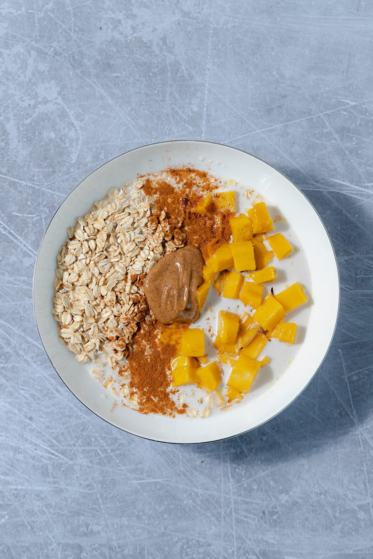 all ingredients for mango overnight oats in a shallow white bowl on a scratched grey metal background.