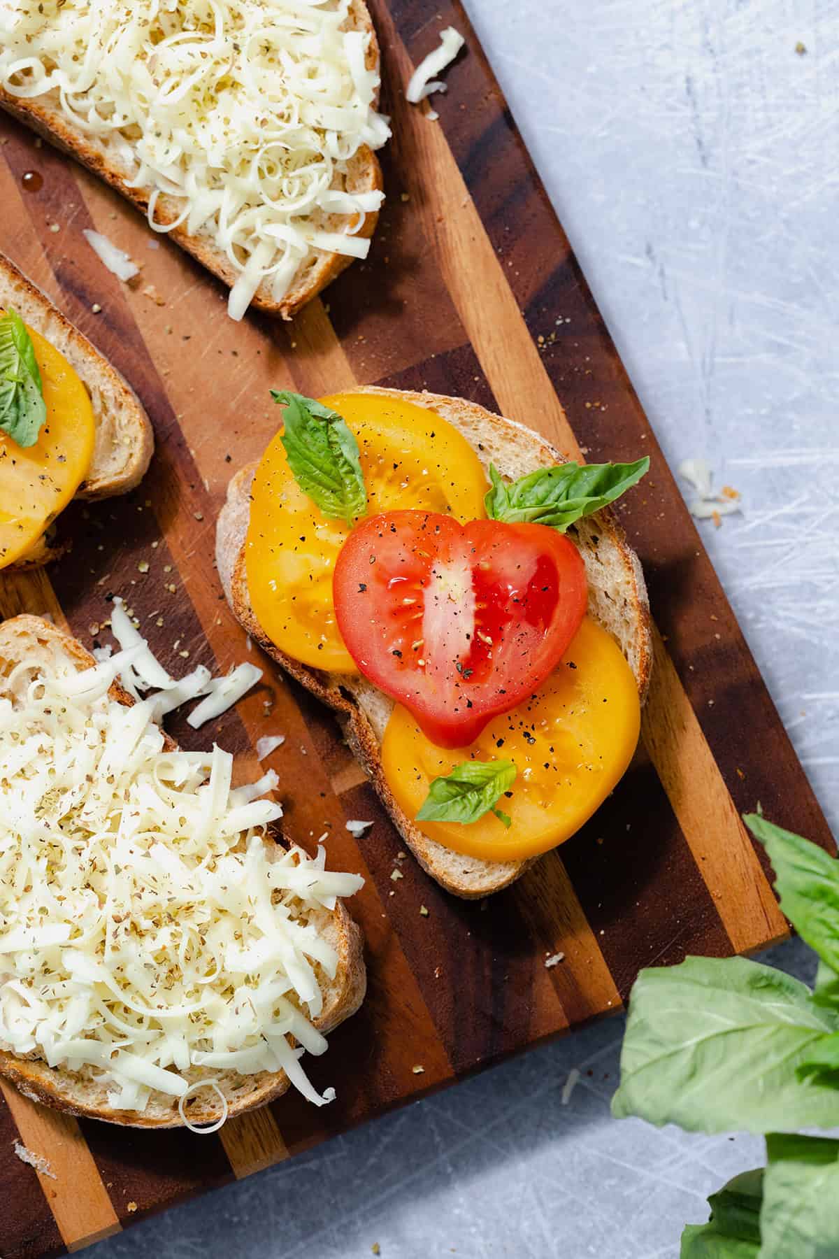 Four slices of bread on a cutting board. The main slice in focus in the middle, layered with sliced heirloom tomatoes and fresh basil. The slice on the bottom left layered with shredded cheese.