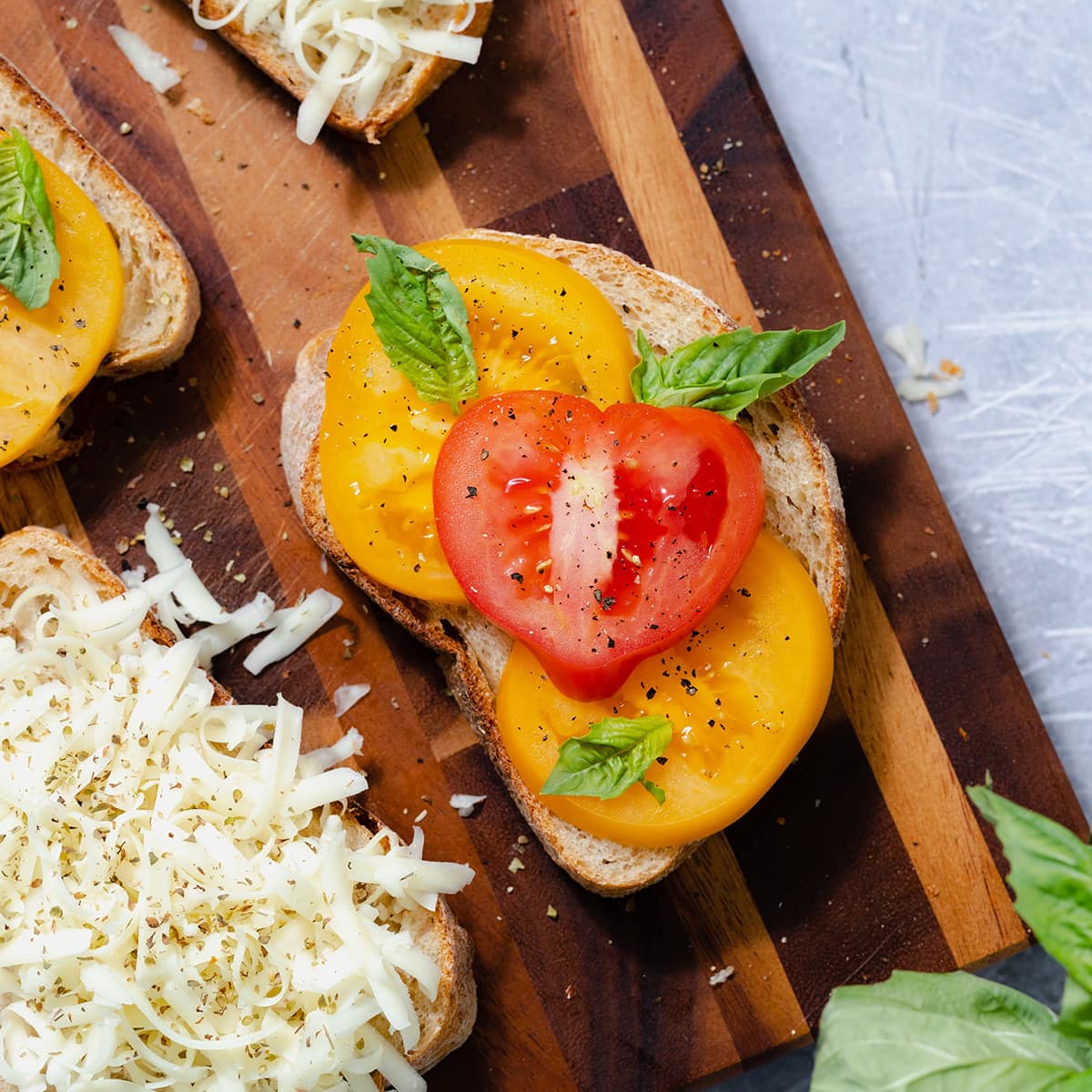 Four slices of bread on a cutting board. The main slice in focus in the middle, layered with sliced heirloom tomatoes and fresh basil. The slice on the bottom left layered with shredded cheese.