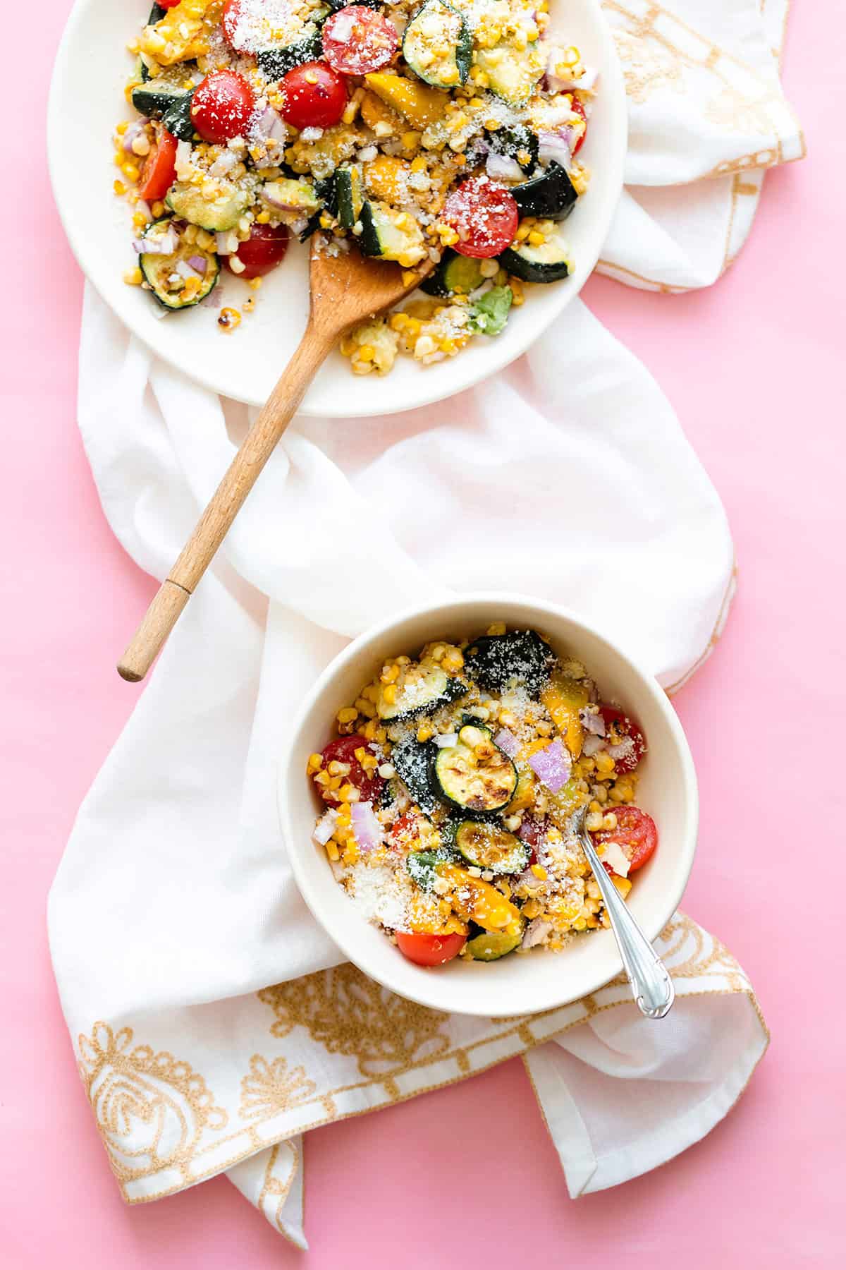 Charred Corn Zucchini Salad sprinkled with Pecorino Cheese in a small white bowl and a white plate in the top left corner. On a pink background. with a beige tea towel under the bowl.