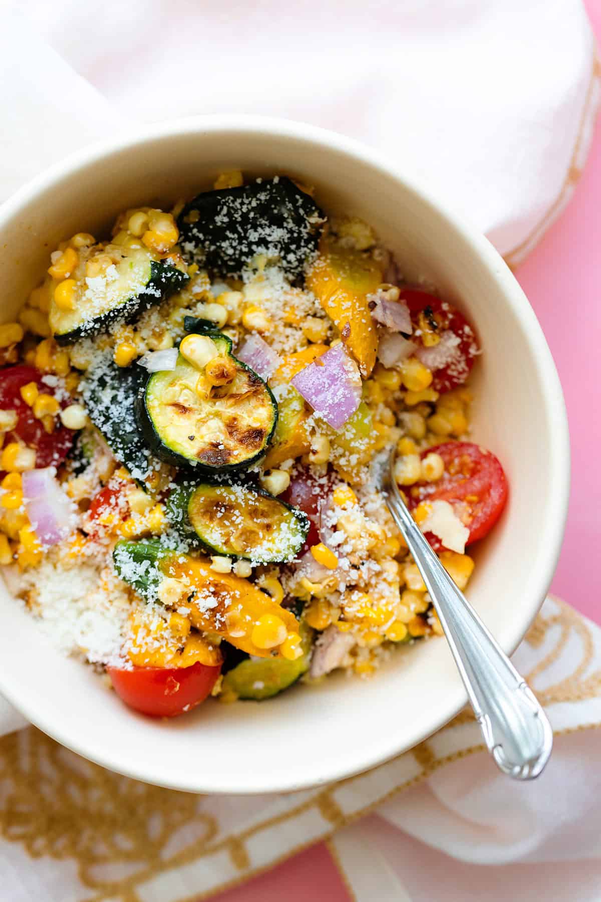 Charred Corn Zucchini Salad sprinkled with Pecorino Cheese in a small white bowl on a pink background. with a beige tea towel under the bowl.