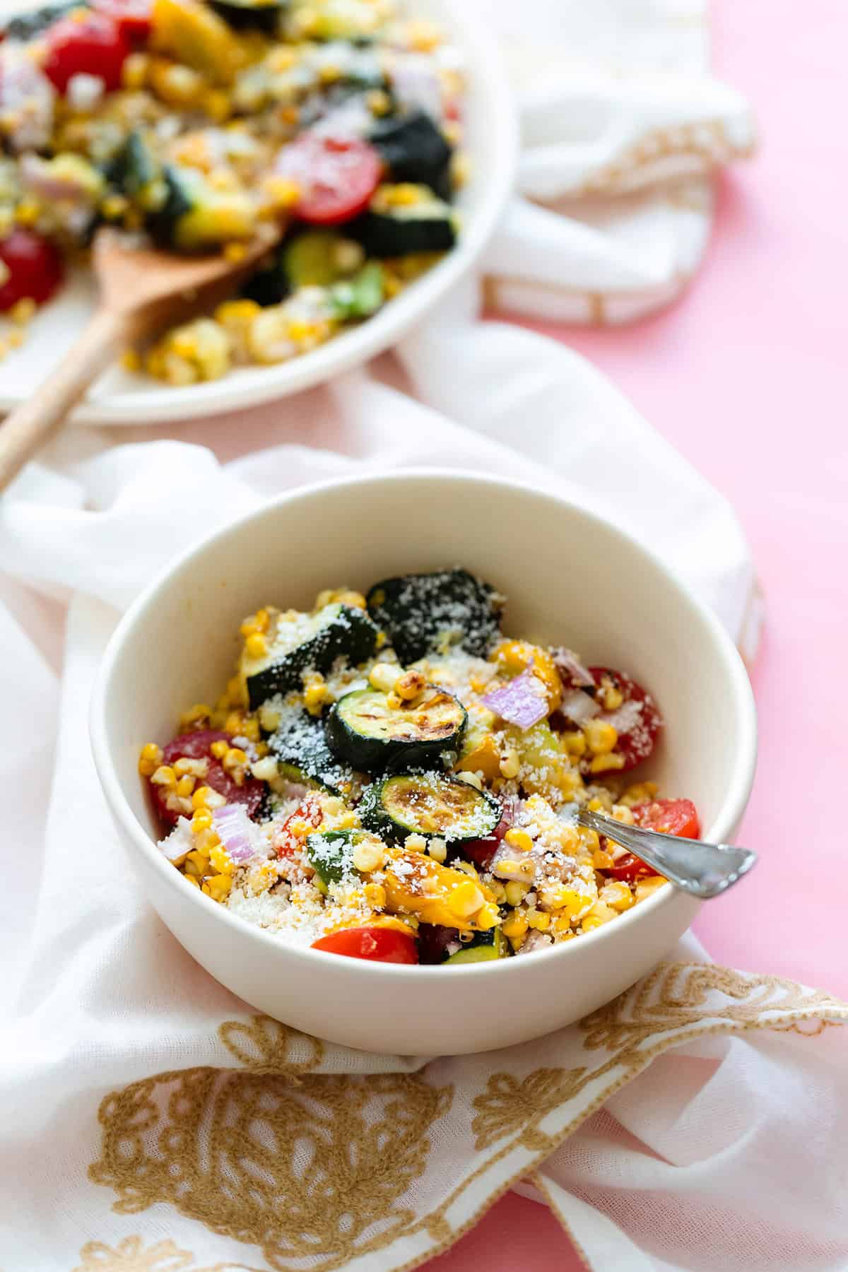 Charred Corn Zucchini Salad sprinkled with Pecorino Cheese in a small white bowl and a white plate in the top left corner. On a pink background. with a beige tea towel under the bowl.