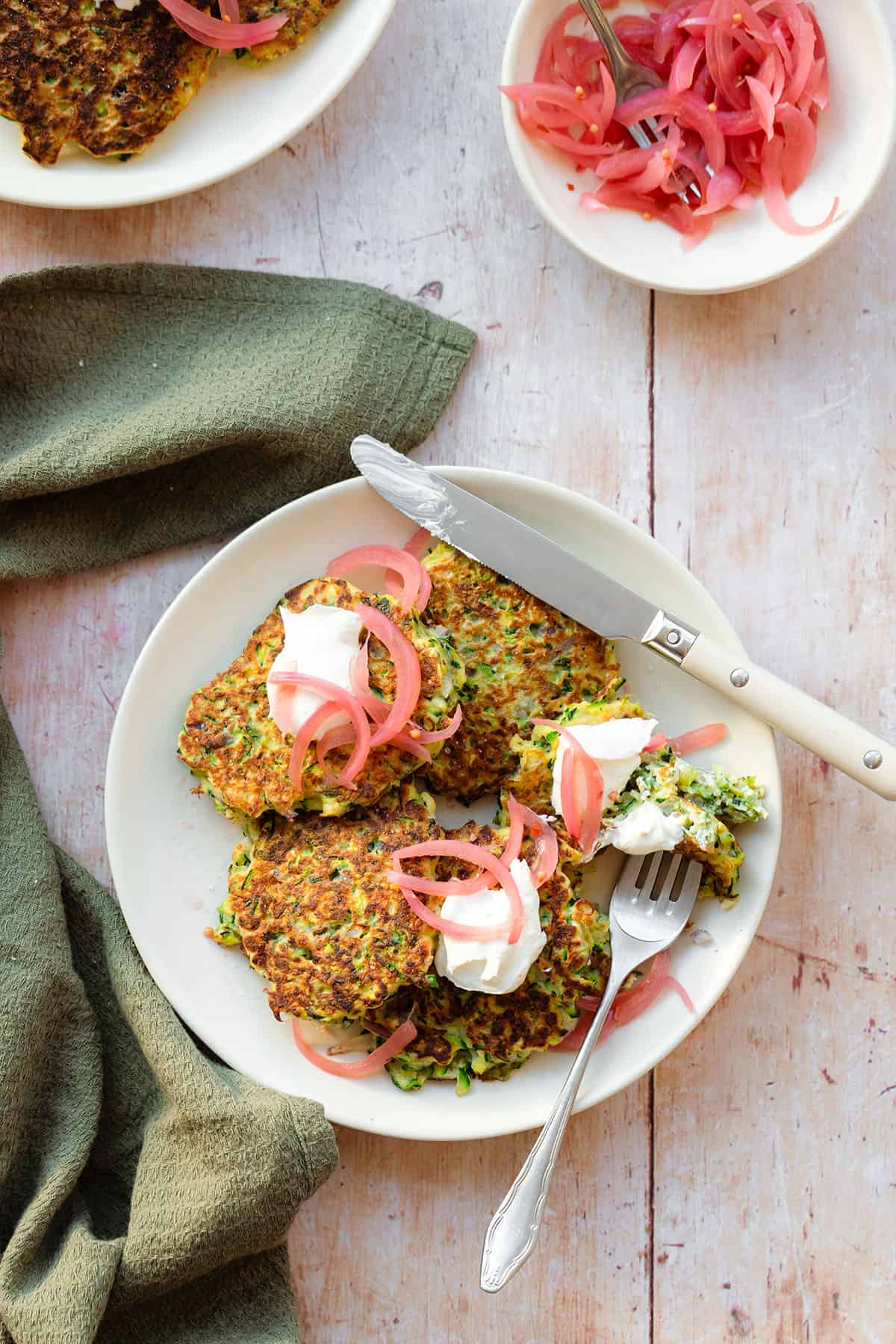 Zucchini fritters topped with pickled red onion and labneh on a white plate on a light wooden background with a green kitchen towel lying on the left. There is a fork and a knife on the plate with a cut up fritter.