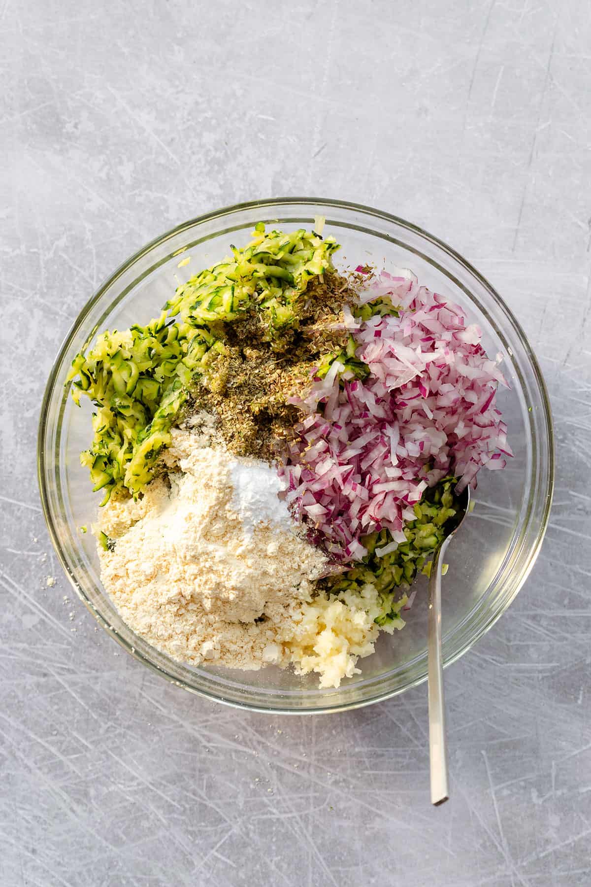 A glass bowl on a scratched steel background. In the bowl, there is grated zucchini chopped red onion, dried marjoram, and chickpea flour.