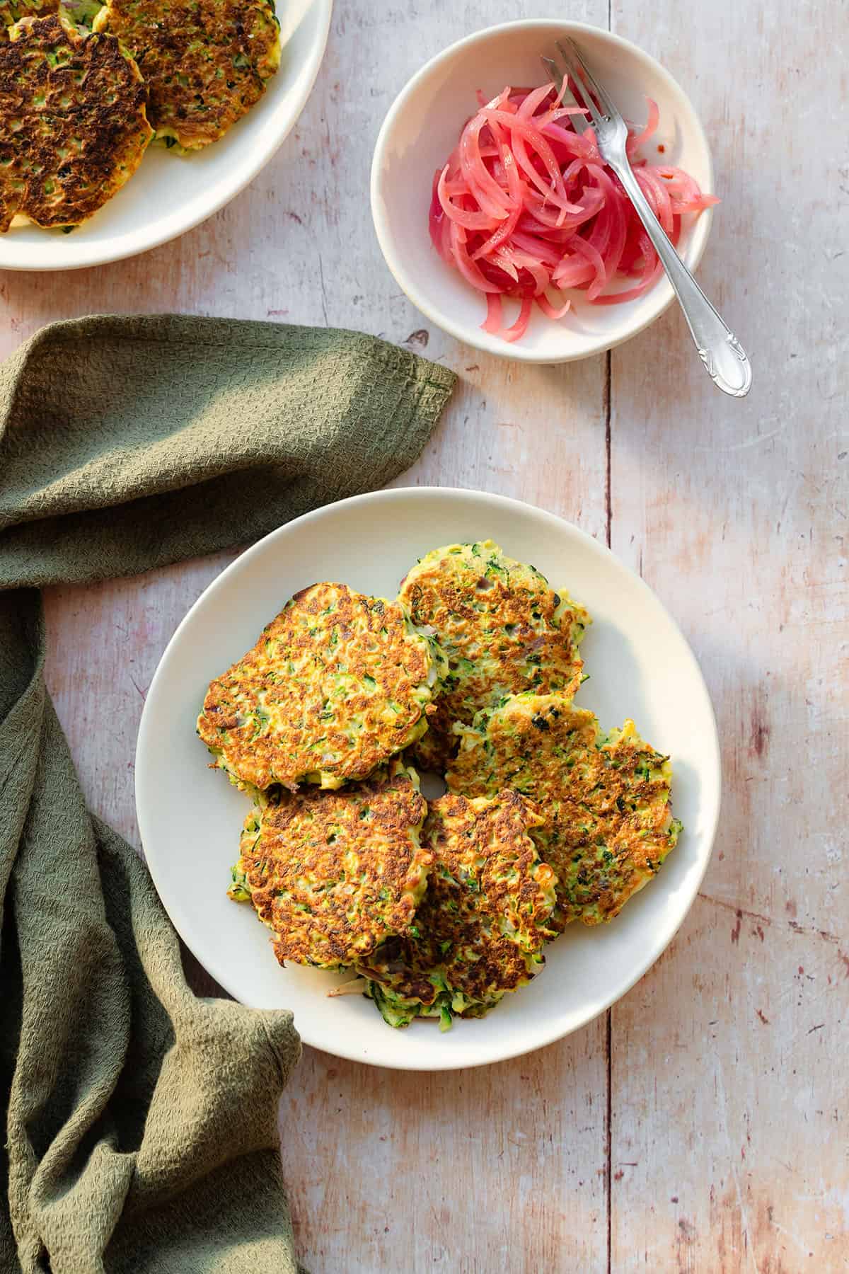 Zucchini fritters on a white plate with pickled red onions served on the side on a small plate on a light wooden background with a green kitchen towel lying on the left and more fritters in the top left corner.