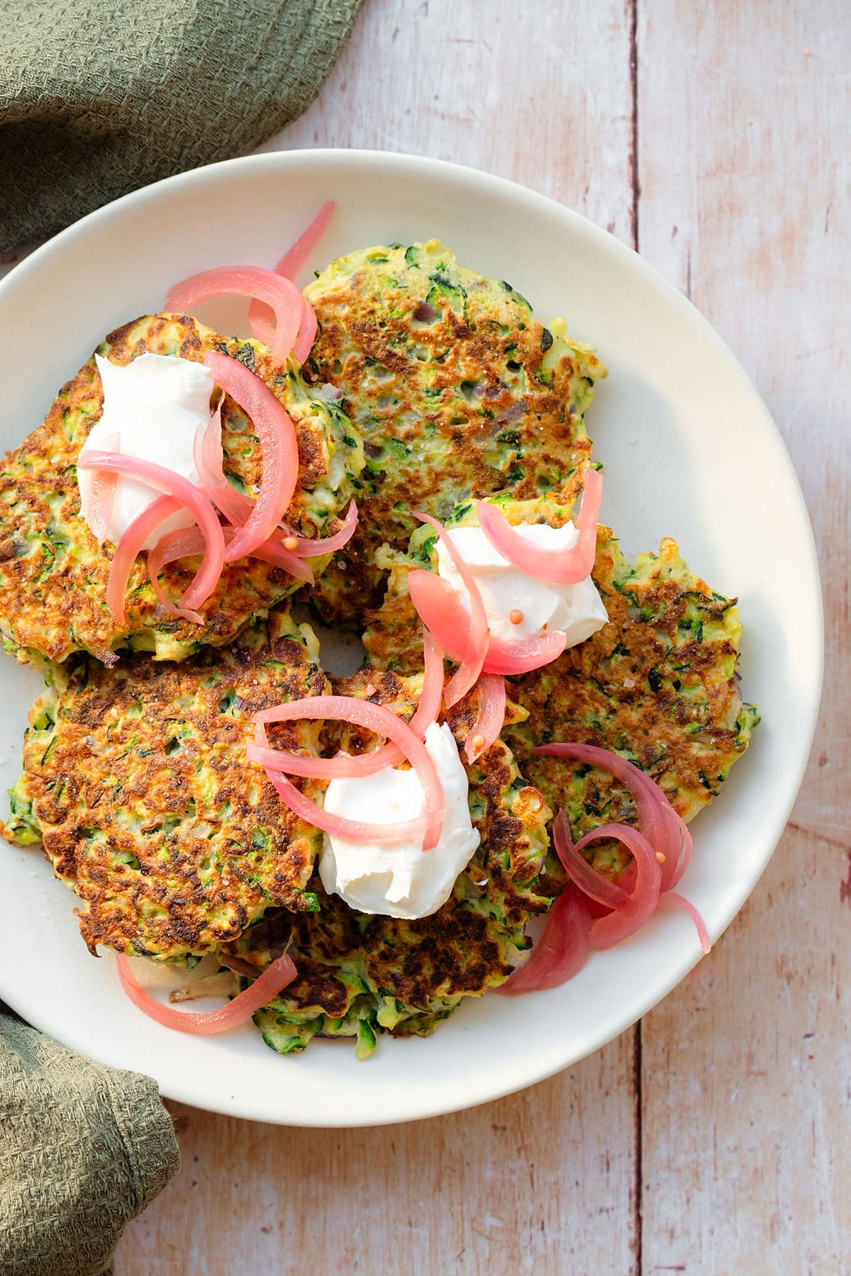 Zucchini fritters topped with pickled red onion and labneh on a white plate on a light wooden background with a green kitchen towel lying on the left.