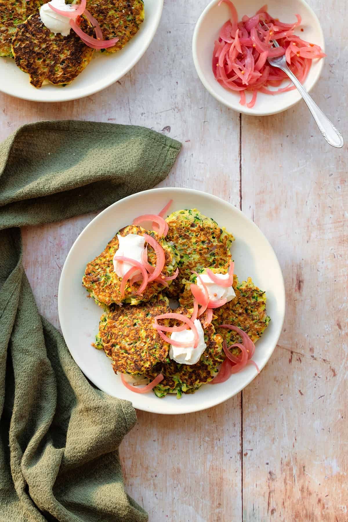 Zucchini fritters topped with pickled red onion and labneh on a white plate on a light wooden background with a green kitchen towel lying on the left.