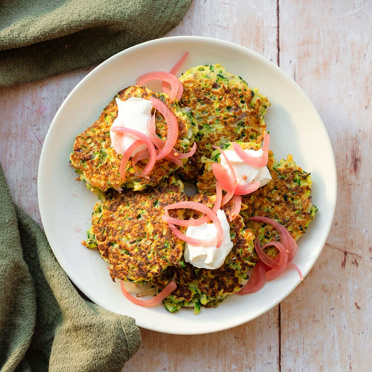 Zucchini fritters topped with pickled red onion and labneh on a white plate on a light wooden background with a green kitchen towel lying on the left.