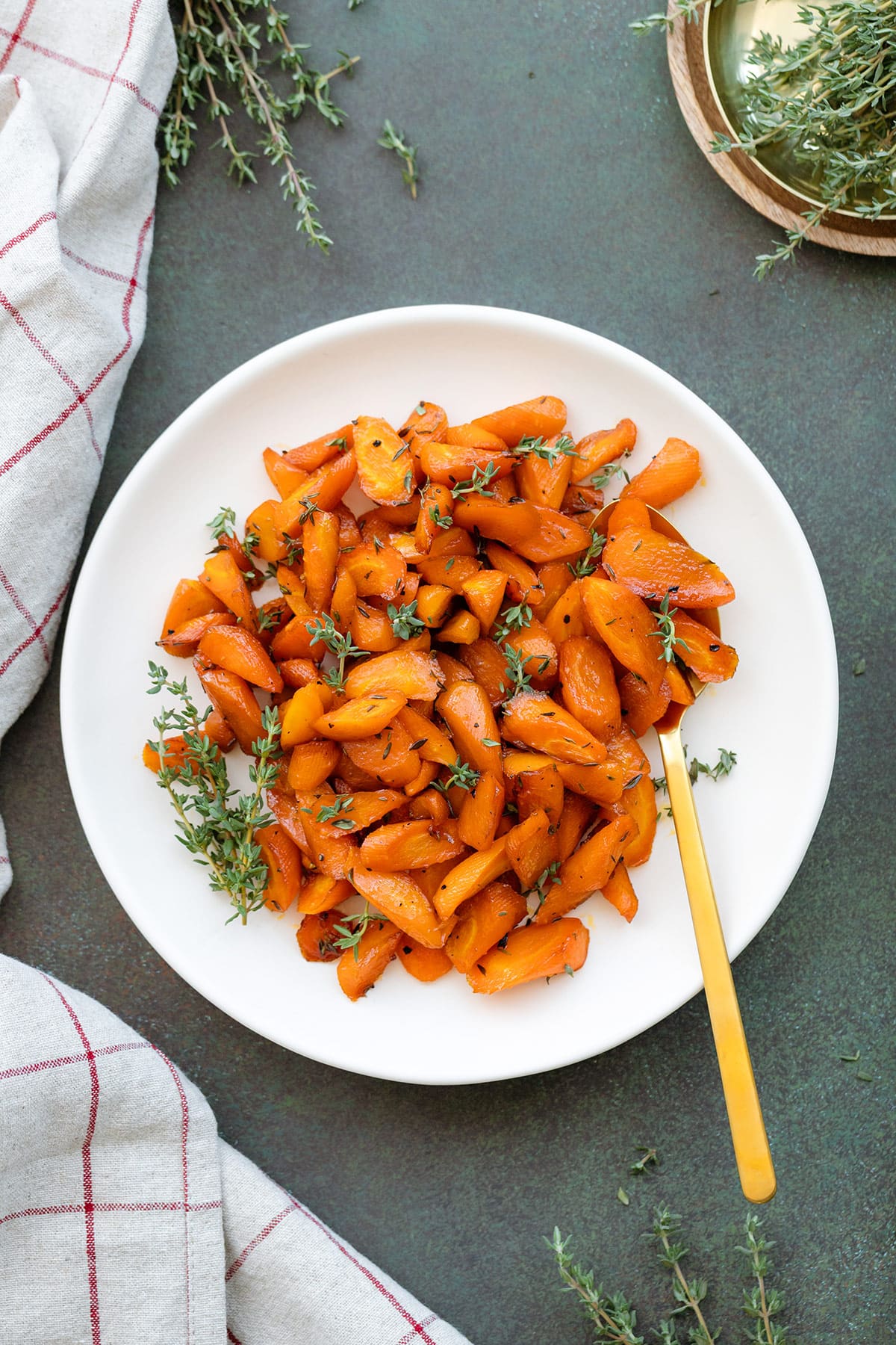 Roasted carrots garnished with fresh thyme on a white plate with a gold spoon on a dark green background.