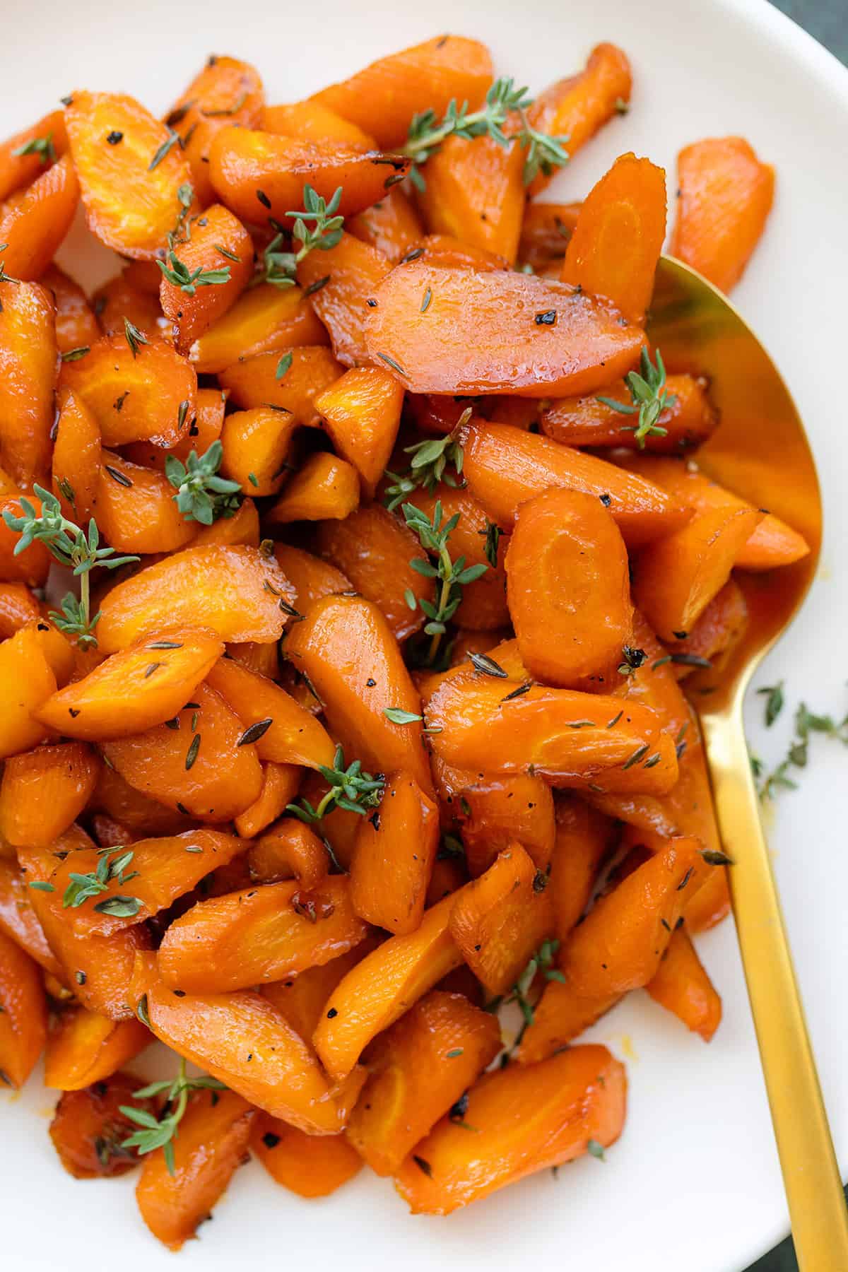 A close up of roasted carrots garnished with fresh thyme on a white plate with a gold spoon on a dark green background.