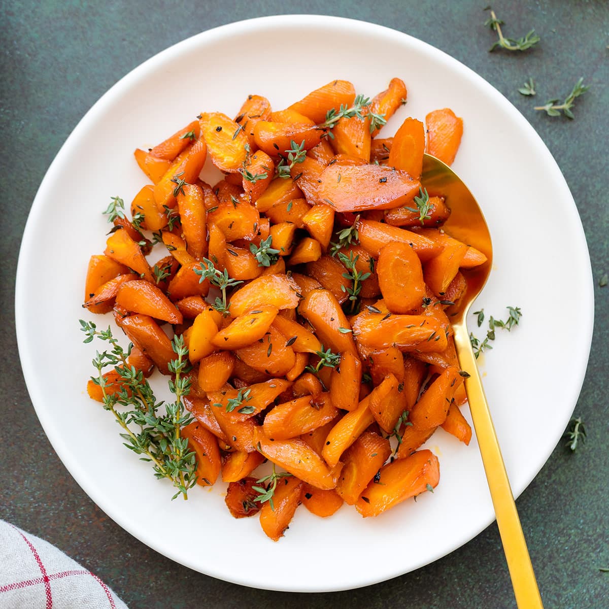 Roasted carrots garnished with fresh thyme on a white plate with a gold spoon on a dark green background.