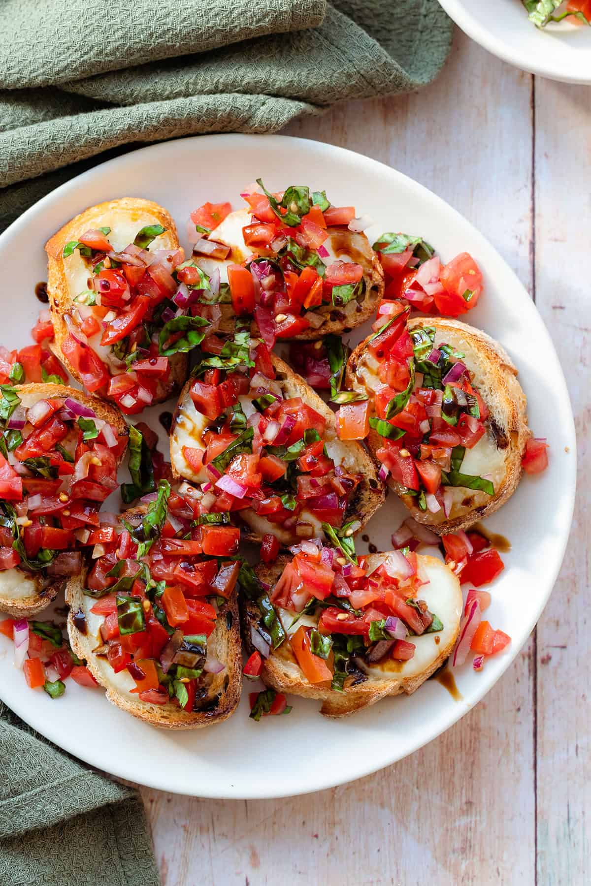 A close up of Bruschetta on a white plate shot from above on a light wooden background with a green kitchen towel on the left.