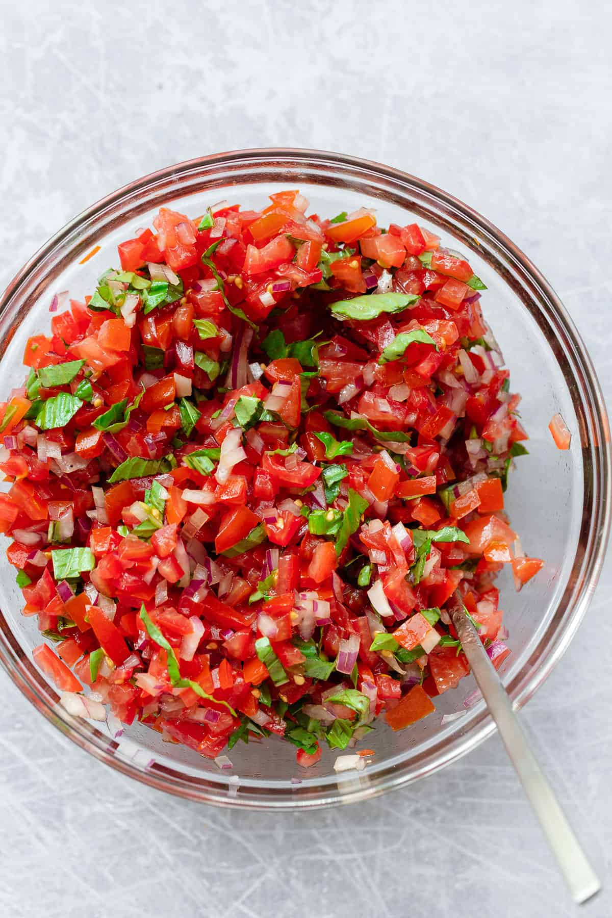 A gladd bowl with a mixture of diced tomatoes, basil, red onion, and balsamic. Shot from above on a scratched metal background.