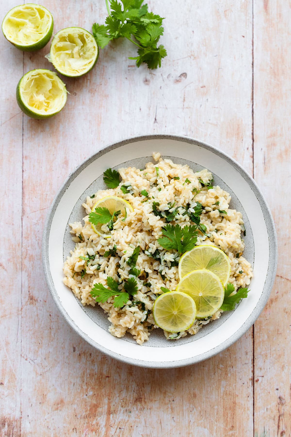 A photo of brown rice with cilantro and lime, decorated with lime slices. In a grey bowl with a white rim on a light wooden background. Juiced lime and fresh cilantro in the left top corner.
