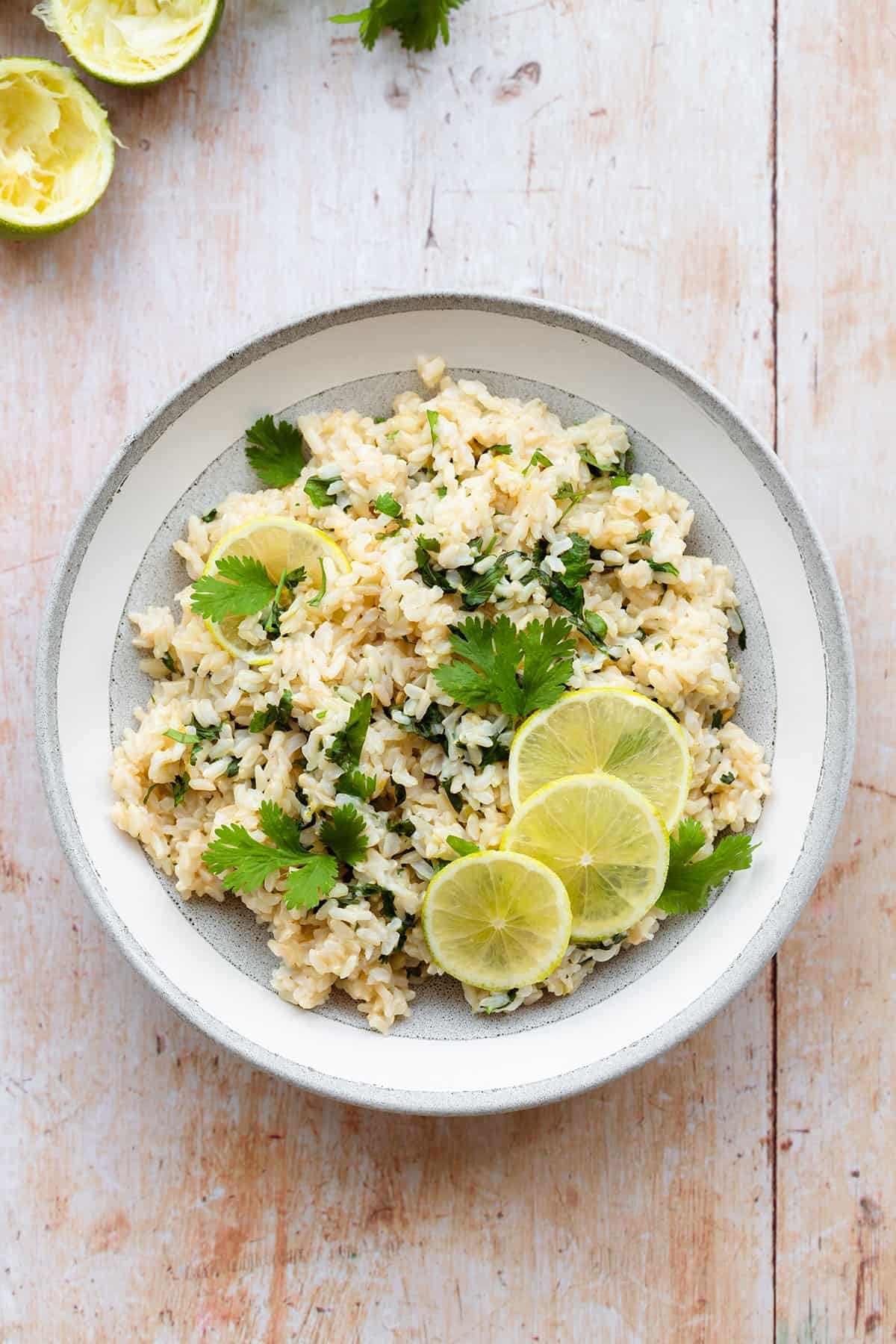 A photo of brown rice with cilantro and lime, decorated with lime slices. In a grey bowl with a white rim on a light wooden background. Juiced lime and fresh cilantro in the left top corner.