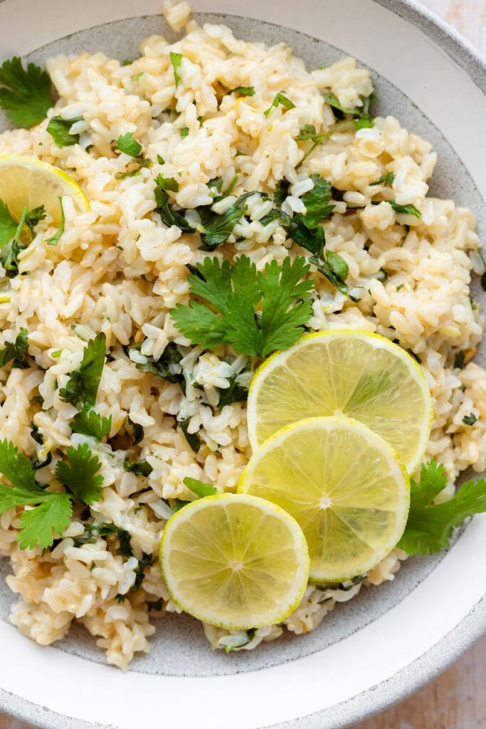 A close up of brown rice with cilantro and lime, decorated with lime slices. In a grey bowl with a white rim on a light wooden background.
