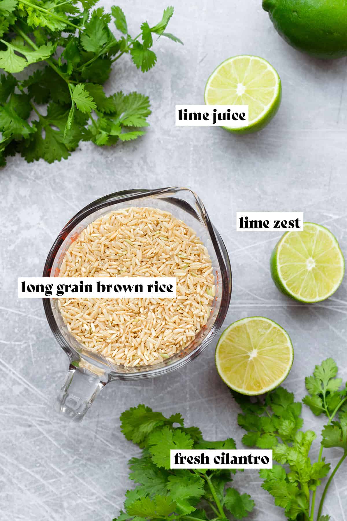 A photo of dry brown long-grain rice in a glass measuring cup with fresh lime and cilantro around on a steel background.