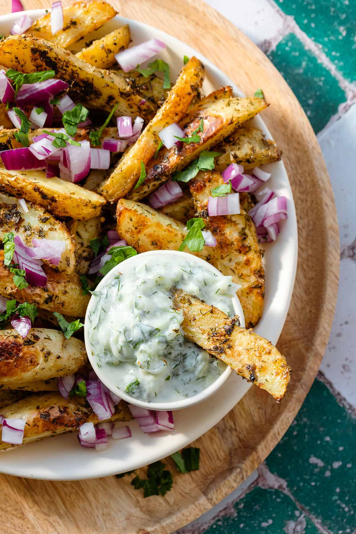 Close up of Greek fries with red onion and parsley on a white plate and a wooden serving plate.