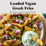 Fries topped with diced red onion, fresh parsley, and tzatziki sauce on the side. One fry is dipped in the sauce. in beige shallow bowl on a wooden serving plate on a green and white tile background. There is a text that says "Loaded Vegan Greek Fries with red onion and parsley".