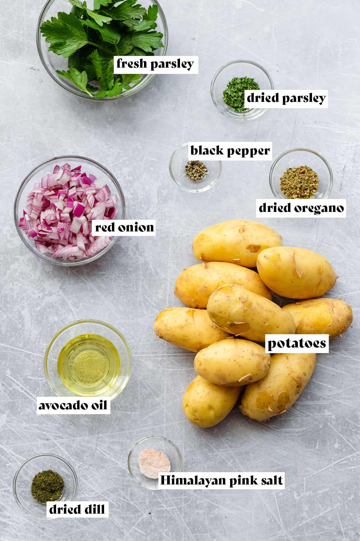 All the ingredients for Greek Fries laid out. There are potatoes, diced red onion, fresh parsley, dried parsley, dried oregano, dried dill, black pepper, Himalayan pink salt, and avocado oil.