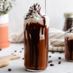 A tall glass with iced mocha topped with whipped cream and a drizzle of dark chocolate on a white background.