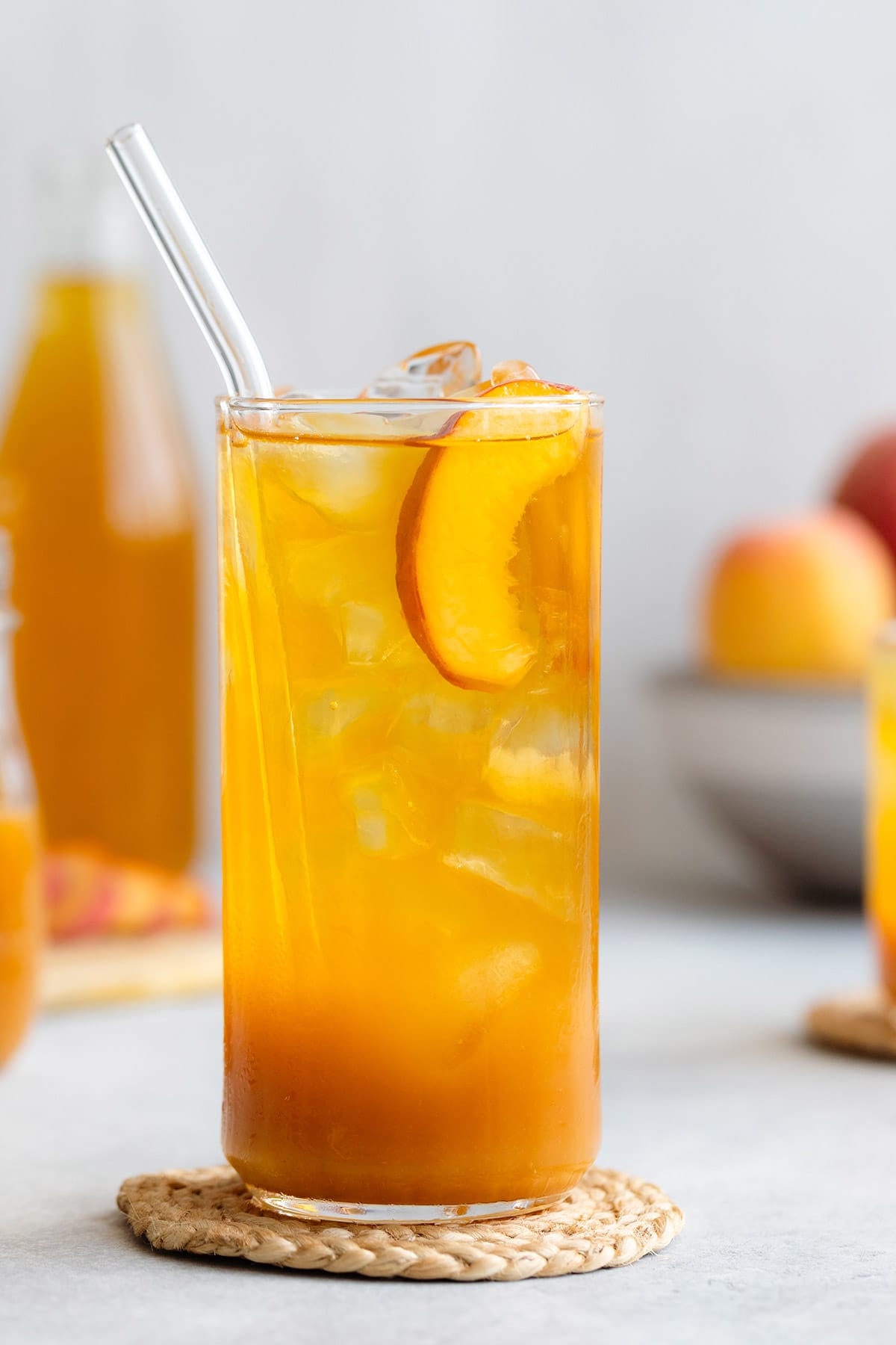 Iced tea in a tall glass with ice and peach slices on a light grey background with more peaches in a bowl in the back.