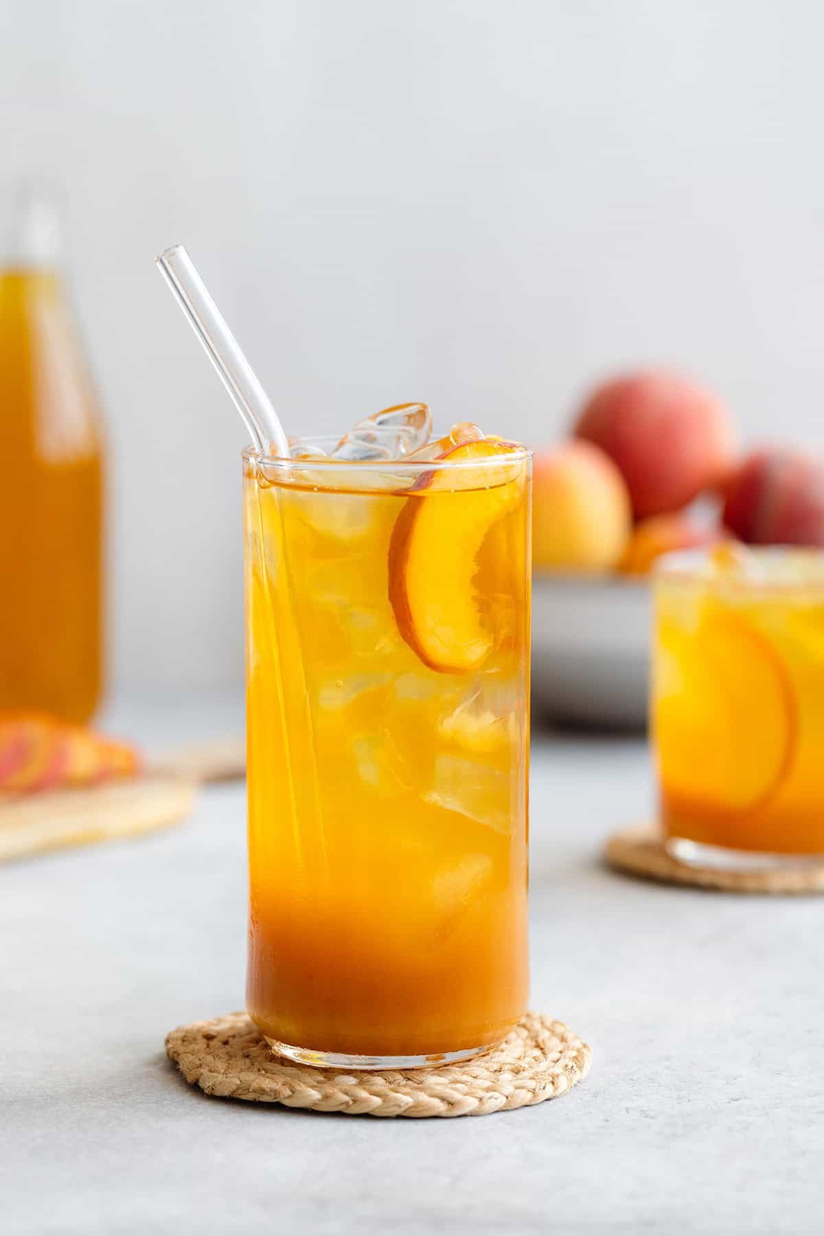 Iced tea in a tall and short glass with ice and peach slices on a light grey background with more peaches in a bowl in the back.