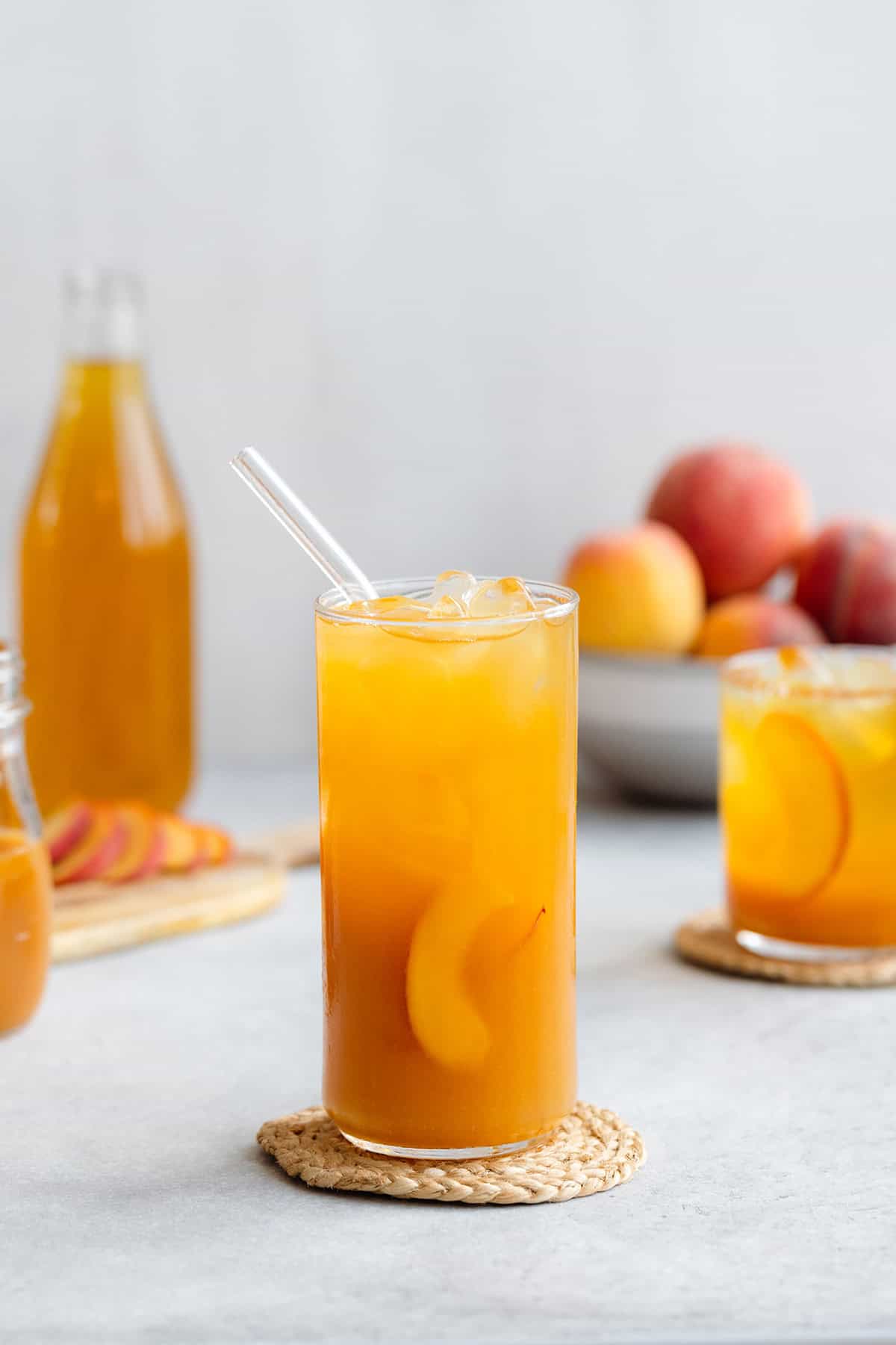 Iced tea in a tall glass with ice and peach slices on a light grey background with more peaches in a bowl in the back.