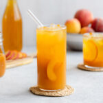 Iced tea in a tall glass with ice and peach slices on a light grey background with more peaches in a bowl in the back.