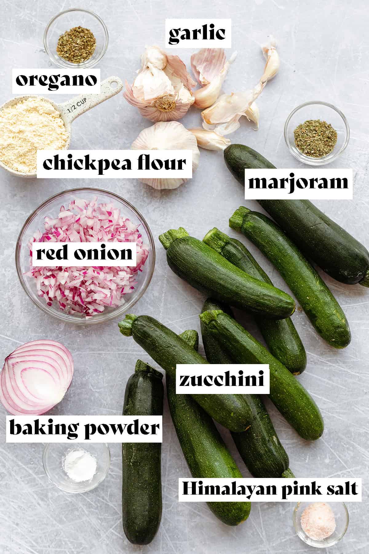 Ingredients to make zucchini fritters layed out on a steel background. Zucchini, red onion, baking powder, himalayan pink salt, marjoram, chickpea flour, oregano, and garlic.