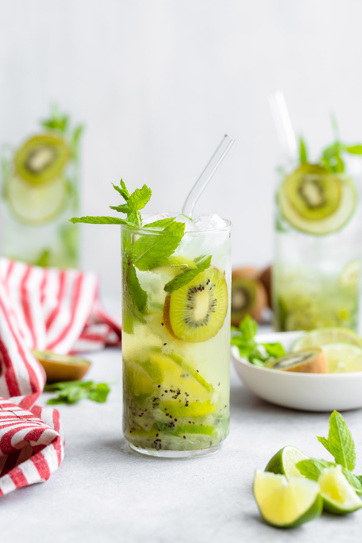 Three Kiwi Mojitos in tall glasses garnished with lime and kiwi slices. On grey background with a striped red and white kitchen towel on the left.