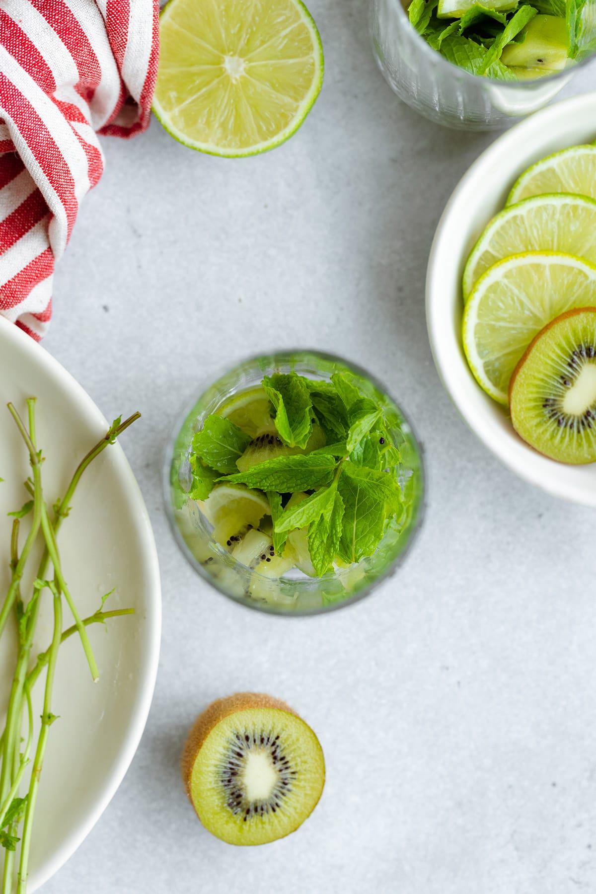 Kiwi, mint, and lime in a tall glass with more fruit all around. Shot from above.