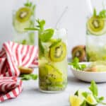 Three Kiwi Mojitos in tall glasses garnished with lime and kiwi slices. On grey background with a striped red and white kitchen towel on the left.