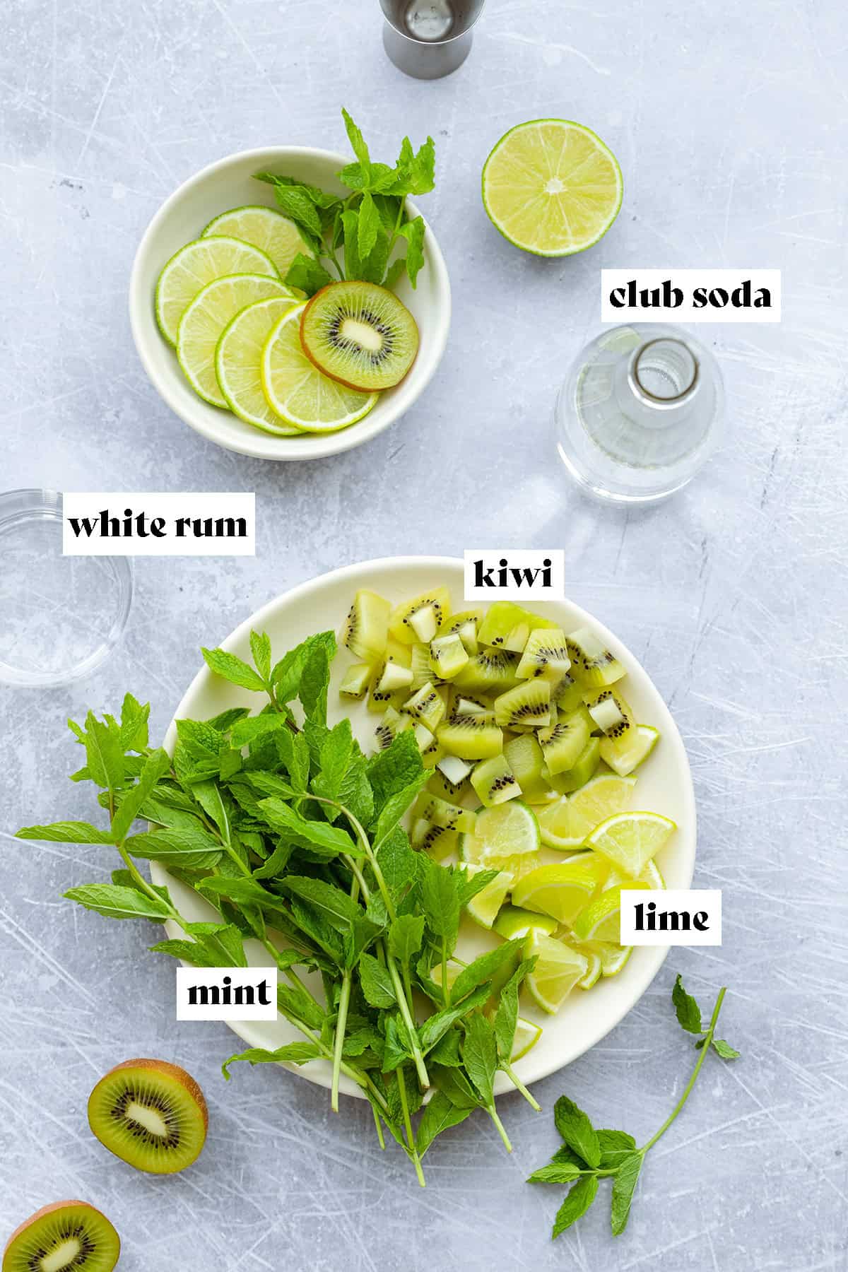 Ingredients for the cocktail laid out on a scratched metal background. There is chopped kiwi, lime, fresh mint, club soda, and white rum.