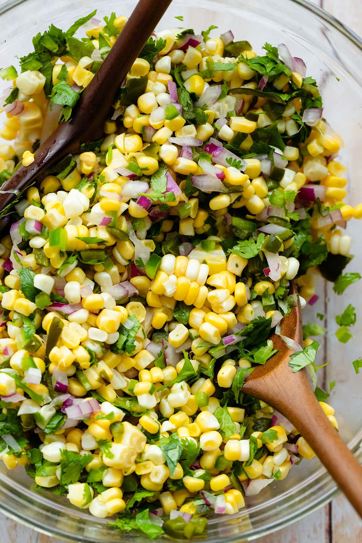 A close up of a glass bowl with Roasted Chili Corn salsa with two wooden mixing spoons on a light wooden backgrond.