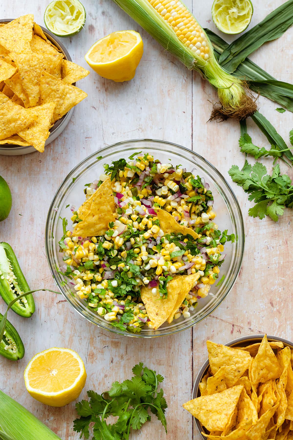 Corn salsa in a glass bowl with tortilla chips dipped in. There are more tortilla chips, fresh cilantro, corn, and jalapenos around. All on a light wooden background.